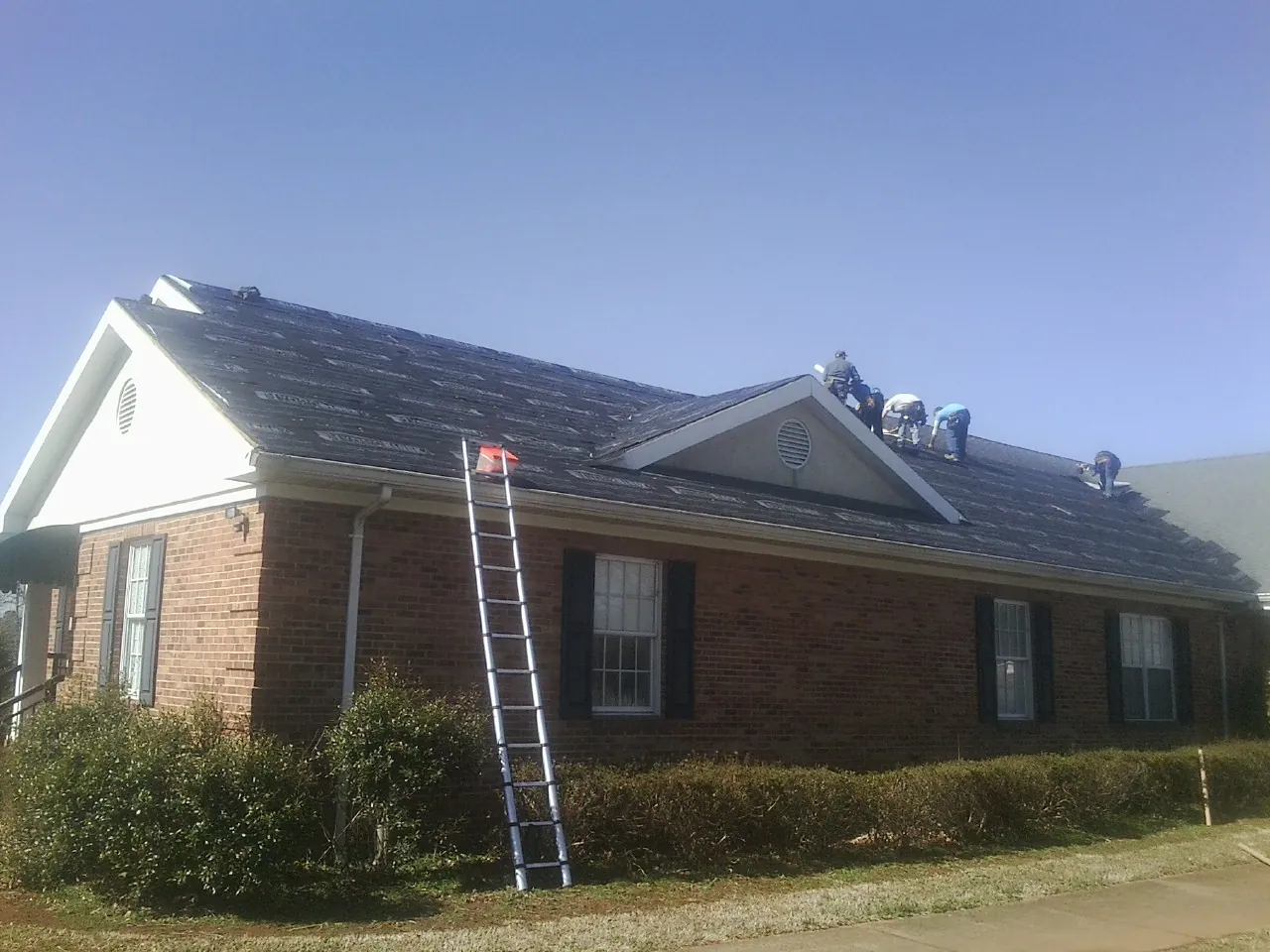 Crew tearing shingles off a brick ranch roof with a ladder set — Metal Roof Repair work by our crew in Nancy