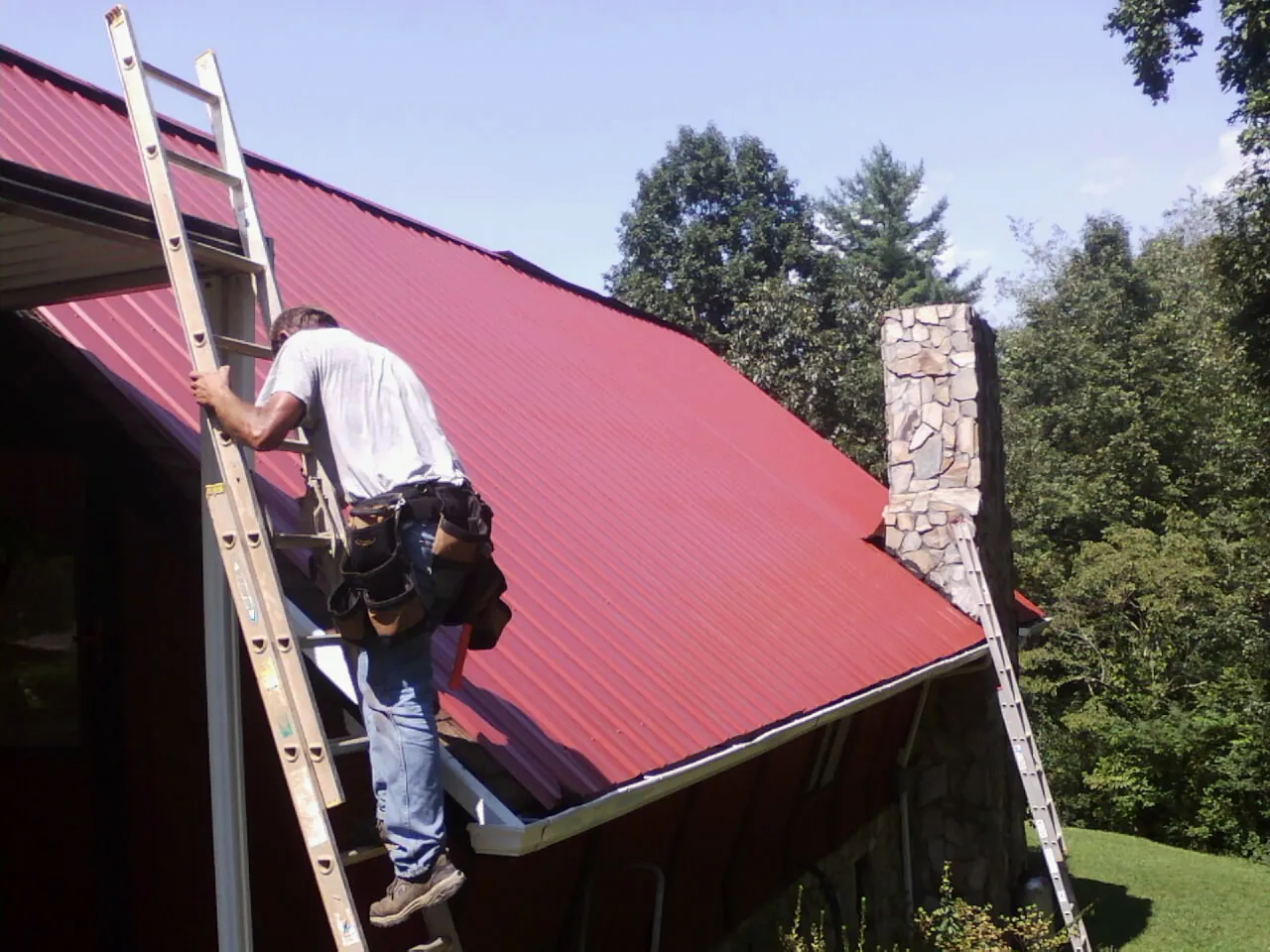Roofer on a ladder beside a red metal roof and stone chimney — Soffit & Fascia Repair by Roofing Brothers in Nancy